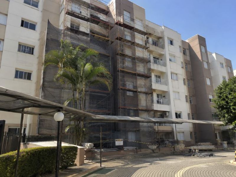 Fachada de prédio residencial em reforma, andaimes metálicos revestidos com tela de proteção instalados na lateral do edifício, palmeiras e paisagismo no pátio interno do condomínio, dia ensolarado com céu azul, fotografia realista de obra de reforma predial